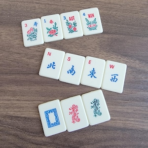 以下が含まれることがあります： 中国の文字とシンボルが書かれた麻雀牌のセット。牌は白地に赤と青のアクセントが付けられている。牌は2列に並んでいる。上の列には、数字の3、1、3、4と「寿」、「春」、「AUT」、「WIN」の文字が書かれている。下の列には、「北」、「南」、「東」、「西」の文字と、龍、花、鳥のシンボルが書かれている。