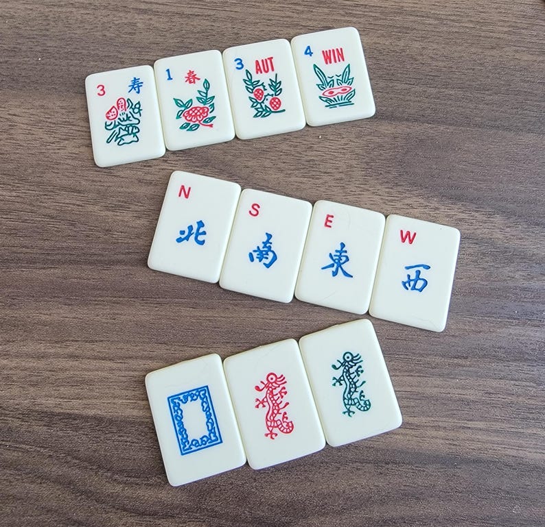 以下が含まれることがあります： 中国の文字とシンボルが書かれた麻雀牌のセット。牌は白地に赤と青のアクセントが付けられている。牌は2列に並んでいる。上の列には、数字の3、1、3、4と「寿」、「春」、「AUT」、「WIN」の文字が書かれている。下の列には、「北」、「南」、「東」、「西」の文字と、龍、花、鳥のシンボルが書かれている。
