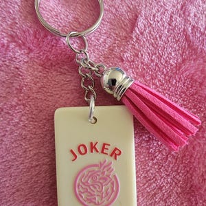 May include: A keychain featuring a rectangular cream-colored charm with the word "JOKER" in red and a pink circular design. It includes a pink tassel and a silver keyring.