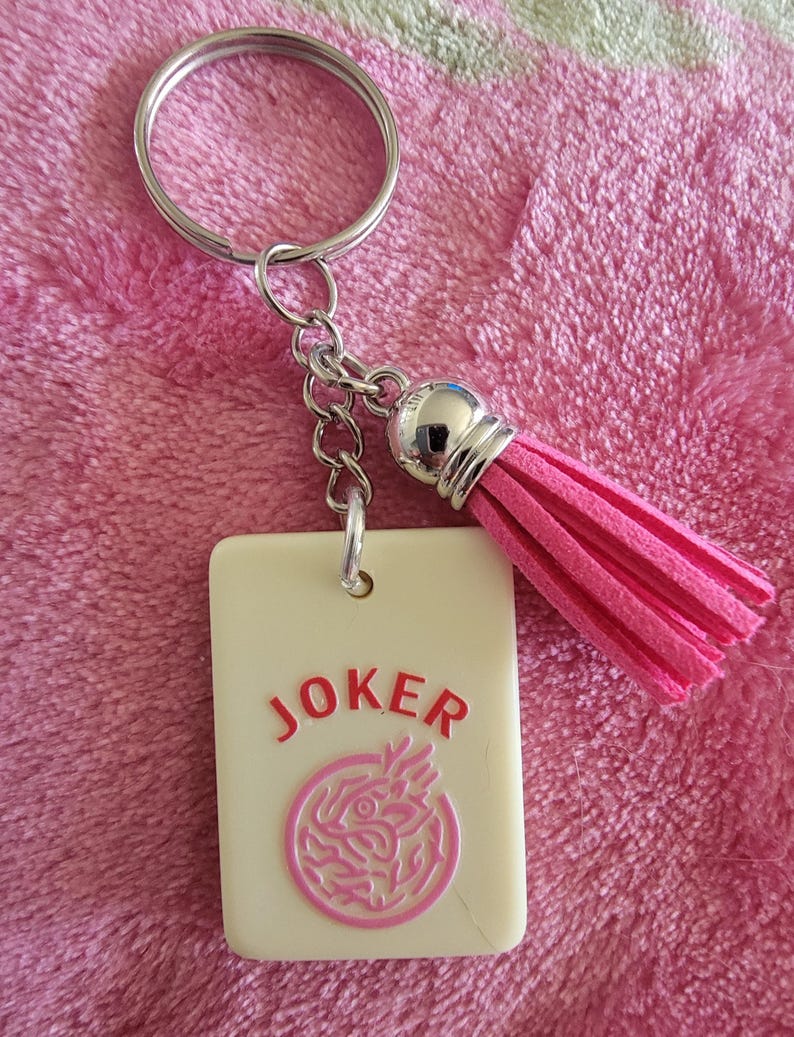 May include: A keychain featuring a rectangular cream-colored charm with the word "JOKER" in red and a pink circular design. It includes a pink tassel and a silver keyring.