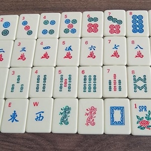 May include: A set of off-white Mahjong tiles arranged in four rows. The tiles feature various colorful engraved designs including numbers, Chinese characters, directional letters N, S, E, W, dragons, a frame, and seasonal symbols labeled 1 SPR and 2 SUM.