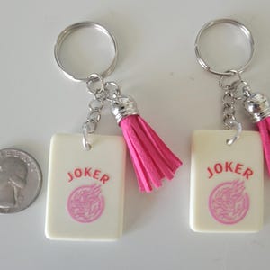 May include: Two keychains with silver rings, chains, and pink tassels. Each keychain has a rectangular cream-colored tag with the word "JOKER" in red and a pink circular design. A quarter is next to the keychains.