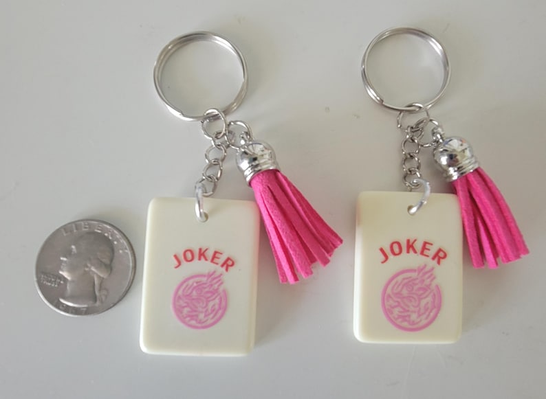 May include: Two keychains with silver rings, chains, and pink tassels. Each keychain has a rectangular cream-colored tag with the word "JOKER" in red and a pink circular design. A quarter is next to the keychains.