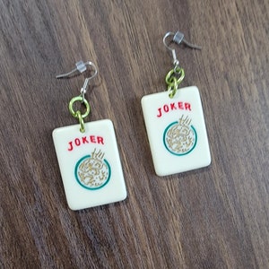May include: A pair of white, rectangular earrings with a green and gold design. The earrings feature the word "JOKER" in red letters and a stylized image of a joker card. The earrings are attached to silver hooks with green and silver chain links.