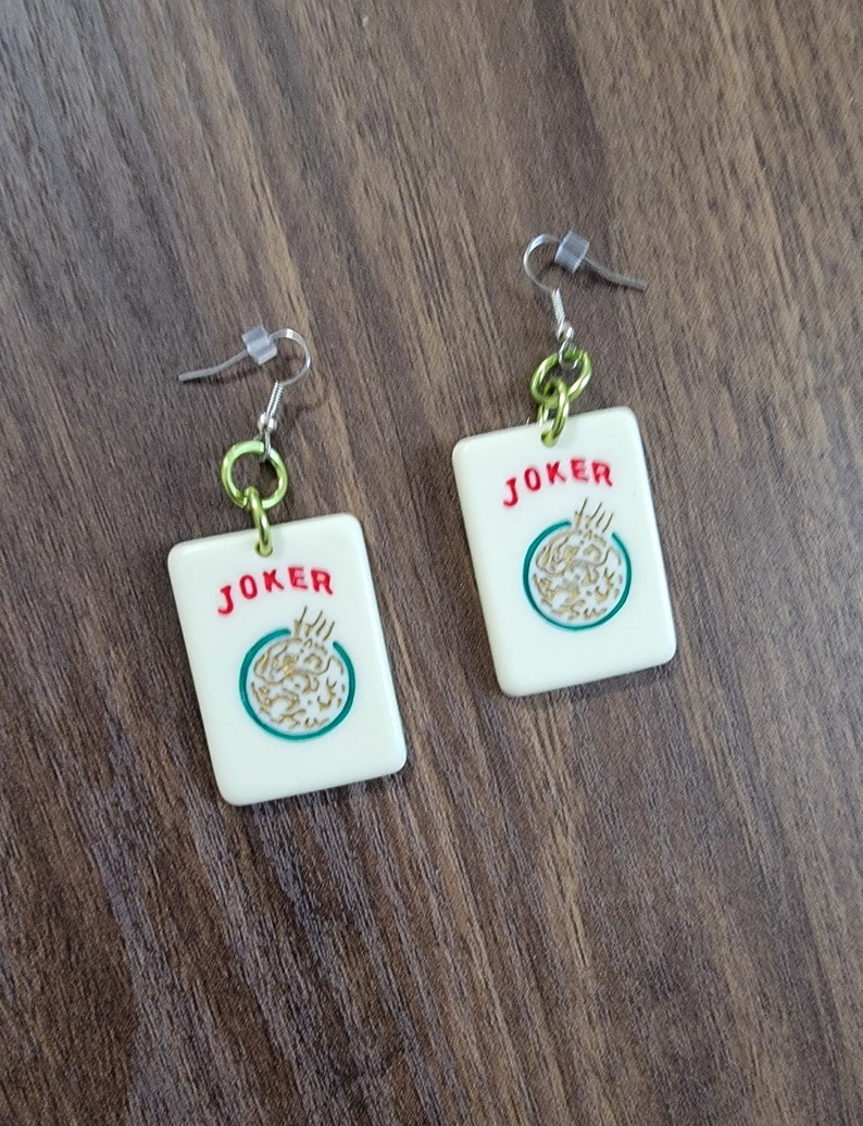 May include: A pair of white, rectangular earrings with a green and gold design. The earrings feature the word "JOKER" in red letters and a stylized image of a joker card. The earrings are attached to silver hooks with green and silver chain links.