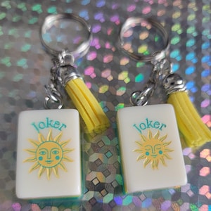 May include: Two keyrings with white rectangular charms featuring a sun design and the word "Joker" in teal. Each keyring has a silver ring, chain, and a yellow tassel. The background is a shiny, iridescent surface.