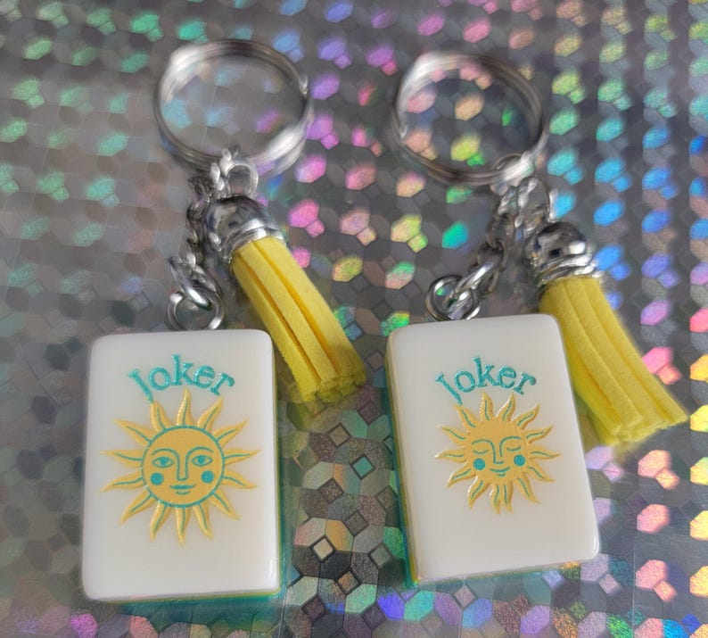 May include: Two keyrings with white rectangular charms featuring a sun design and the word "Joker" in teal. Each keyring has a silver ring, chain, and a yellow tassel. The background is a shiny, iridescent surface.