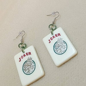 May include: A pair of white, plastic earrings shaped like playing cards. Each earring features a green and white illustration of a joker card with the word "JOKER" printed in red.
