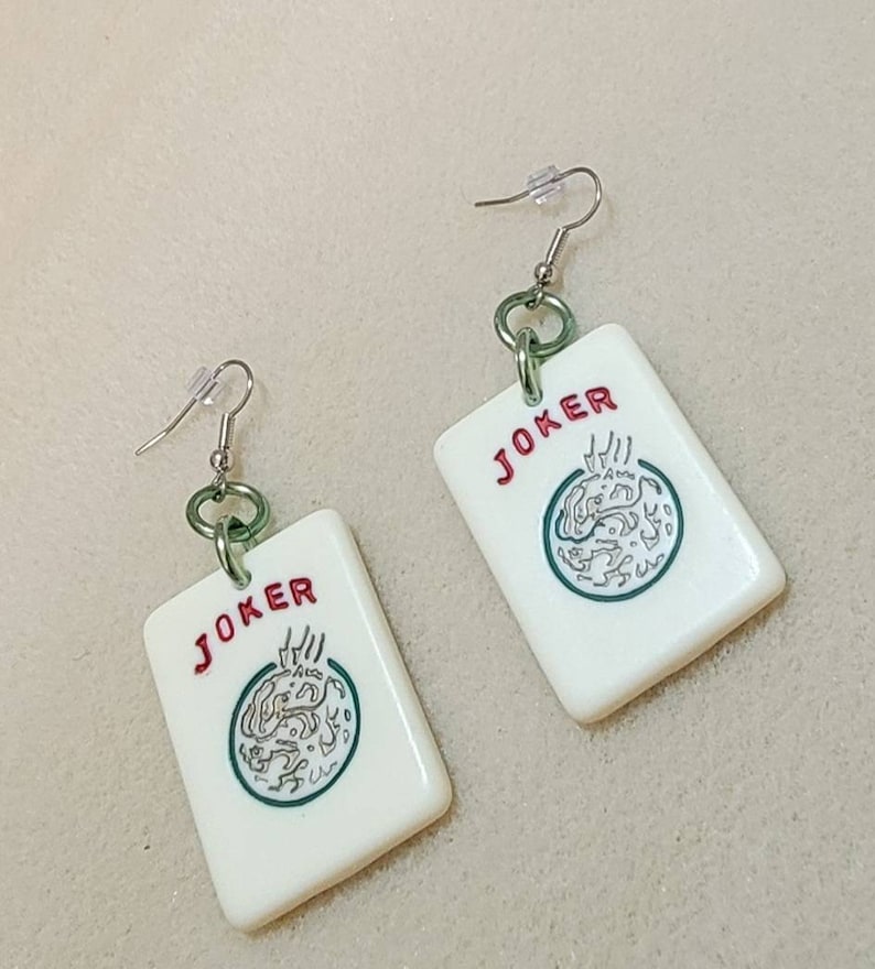 May include: A pair of white, plastic earrings shaped like playing cards. Each earring features a green and white illustration of a joker card with the word "JOKER" printed in red.