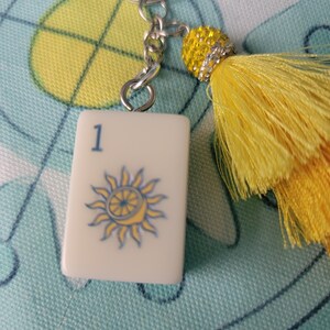 May include: A white tile keychain with a blue number 1 and a sun design. The keychain has a silver chain and a yellow tassel with a beaded accent. The tile is rectangular and has a glossy finish.