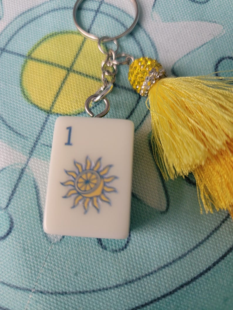 May include: A white tile keychain with a blue number 1 and a sun design. The keychain has a silver chain and a yellow tassel with a beaded accent. The tile is rectangular and has a glossy finish.