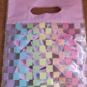 May include: A pink plastic bag with a handle, filled with pink Mahjong tiles. The tiles have various symbols and numbers in different colors. The bag has a woven pattern in pastel colors.