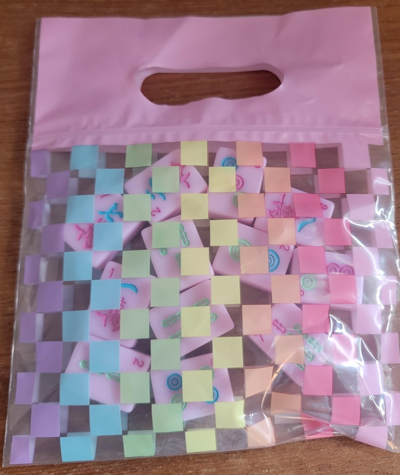 May include: A pink plastic bag with a handle, filled with pink Mahjong tiles. The tiles have various symbols and numbers in different colors. The bag has a woven pattern in pastel colors.