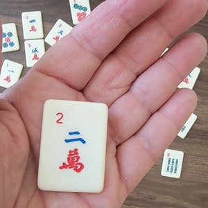 May include: A white tile with a red number 2 and a blue and red Chinese character. The tile is being held in a hand.