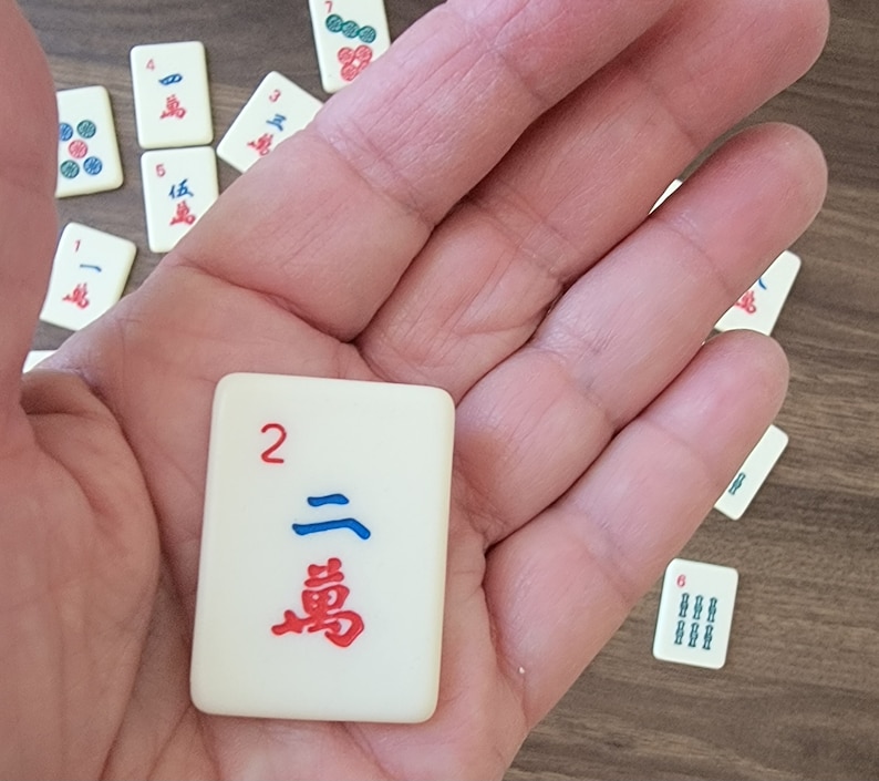 May include: A white tile with a red number 2 and a blue and red Chinese character. The tile is being held in a hand.