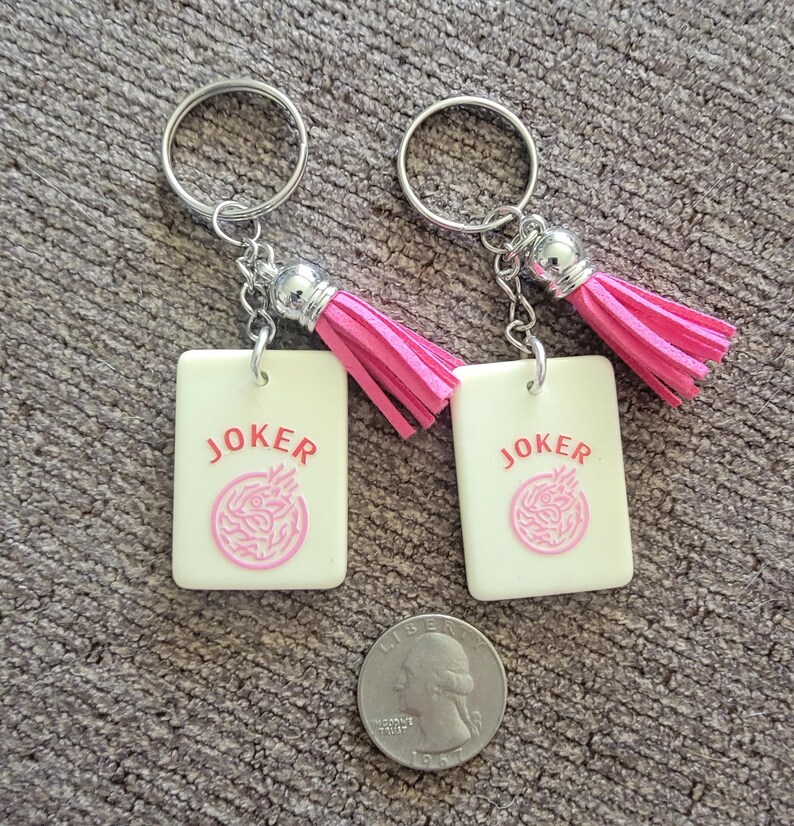 May include: Two keychains with rectangular cream-colored tags. Each tag has the word "JOKER" in red and a pink design. The keychains have silver rings and pink tassels. A quarter is next to the keychains.