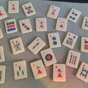May include: A set of 20 mahjong tiles with various numbers, symbols, and characters. The tiles are white with red and blue markings. The tiles are arranged in a random order on a gray surface.
