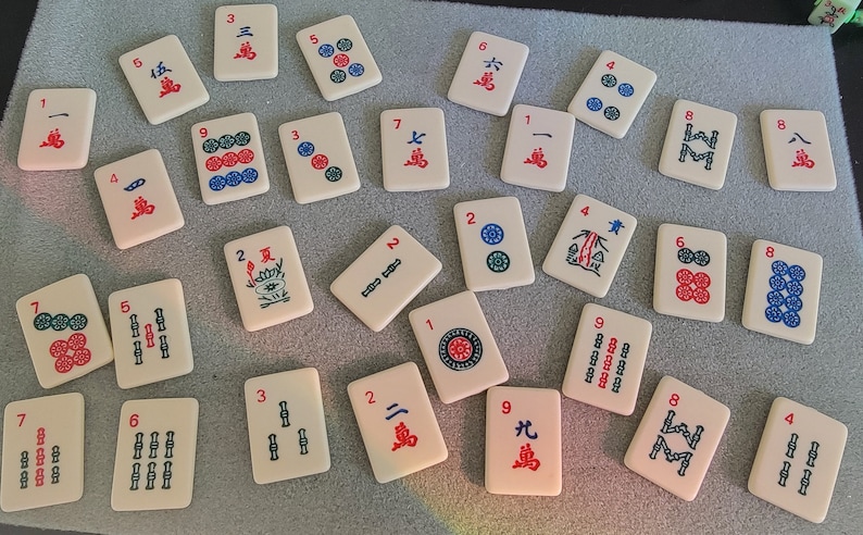 May include: A set of 20 mahjong tiles with various numbers, symbols, and characters. The tiles are white with red and blue markings. The tiles are arranged in a random order on a gray surface.