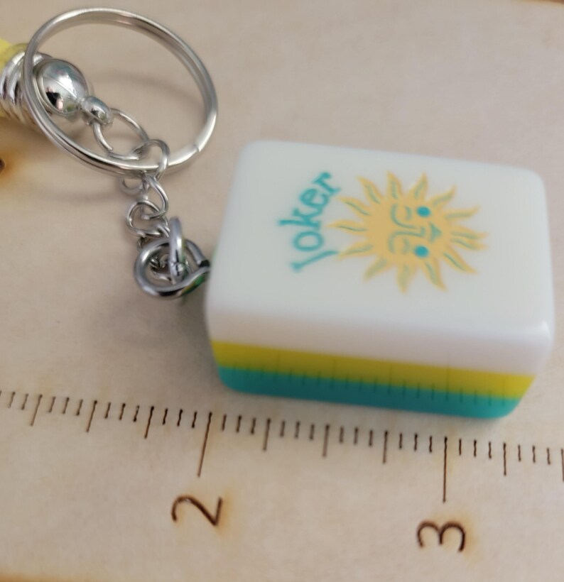 May include: A white and turquoise Mahjong tile keyring with the word "Joker" and a yellow sun design. The keyring has a silver metal ring and a yellow and silver handle.