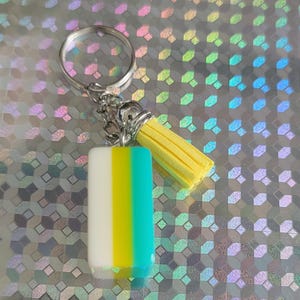 May include: A keychain with a rectangular charm in white, yellow, and teal vertical stripes. It has a yellow tassel and a silver keyring. The background is a holographic, iridescent surface.