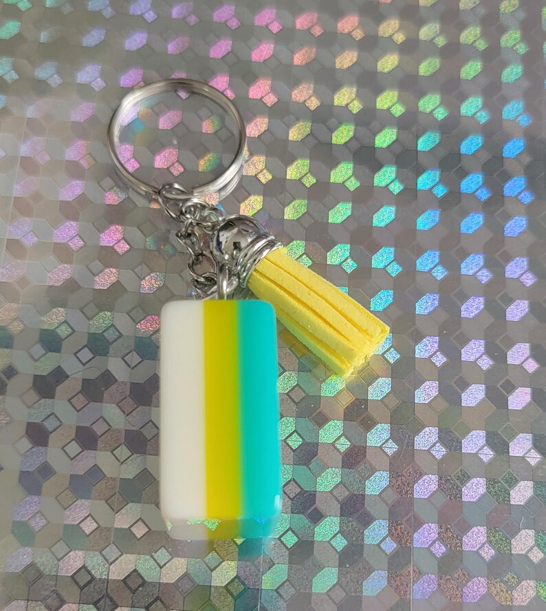 May include: A keychain with a rectangular charm in white, yellow, and teal vertical stripes. It has a yellow tassel and a silver keyring. The background is a holographic, iridescent surface.