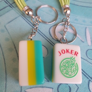 May include: Two keychains with rectangular charms. One charm is white with yellow and teal stripes, the other is white with the word "JOKER" in red and a green dragon design. Each keychain has a silver ring and a green tassel.