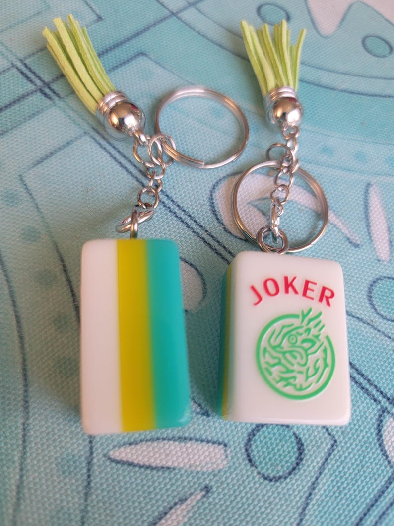 May include: Two keychains with rectangular charms. One charm is white with yellow and teal stripes, the other is white with the word "JOKER" in red and a green dragon design. Each keychain has a silver ring and a green tassel.