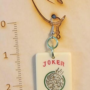 May include: A pair of earrings featuring a white ceramic tile with a green and gold design and the word "JOKER" in red lettering.