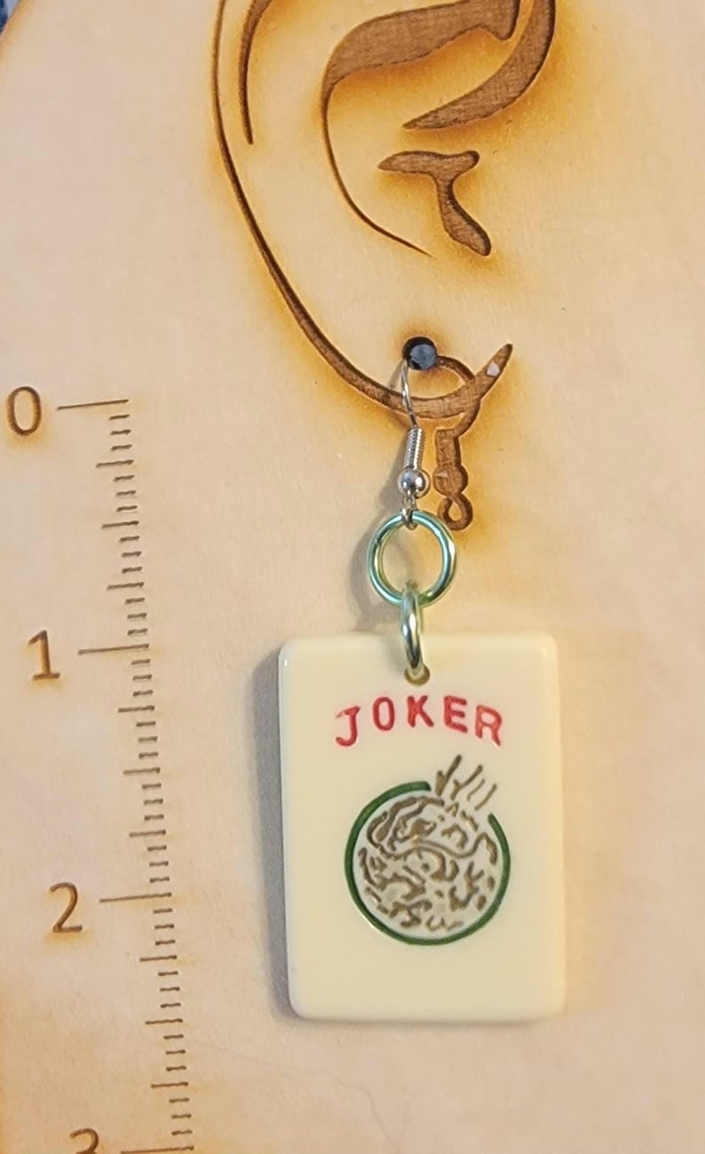 May include: A pair of earrings featuring a white ceramic tile with a green and gold design and the word "JOKER" in red lettering.