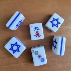 May include: White Mahjong tiles with blue accents and symbols are scattered on a wooden surface. Some tiles feature a blue Star of David, while others display Chinese characters and numbers. The tiles are a classic game accessory.