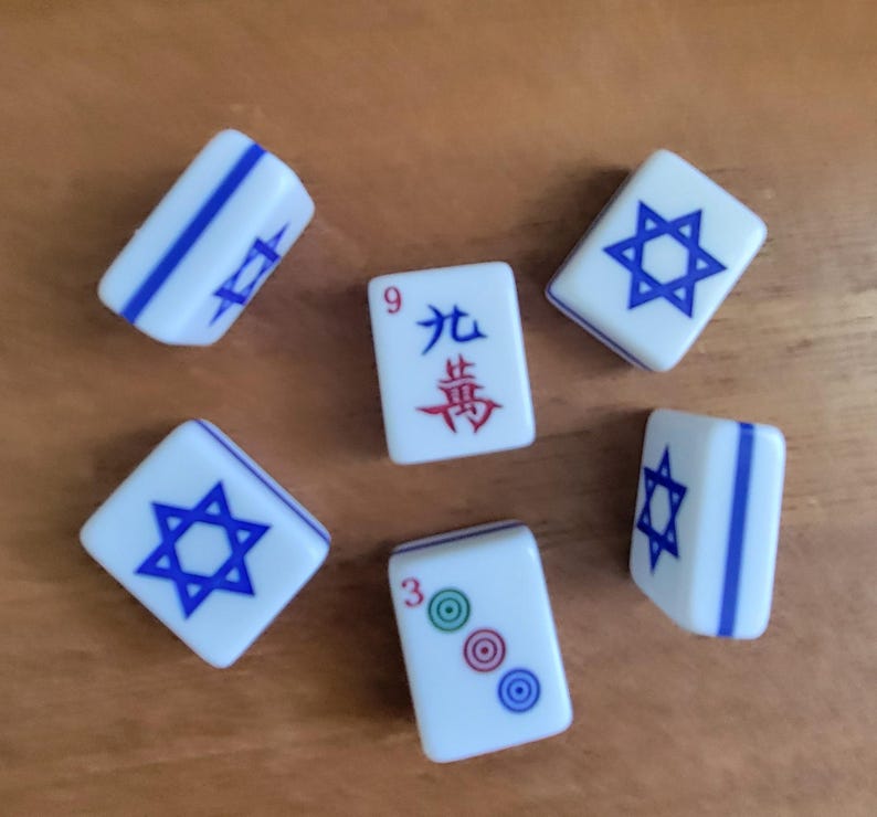 May include: White Mahjong tiles with blue accents and symbols are scattered on a wooden surface. Some tiles feature a blue Star of David, while others display Chinese characters and numbers. The tiles are a classic game accessory.