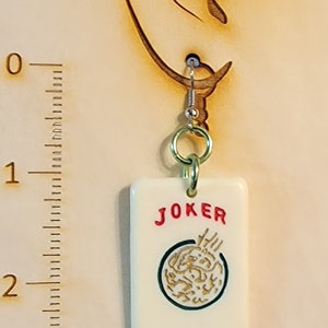 May include: A white ceramic earring with a red "JOKER" inscription and a gold-colored design. The earring is hanging from a silver hook.