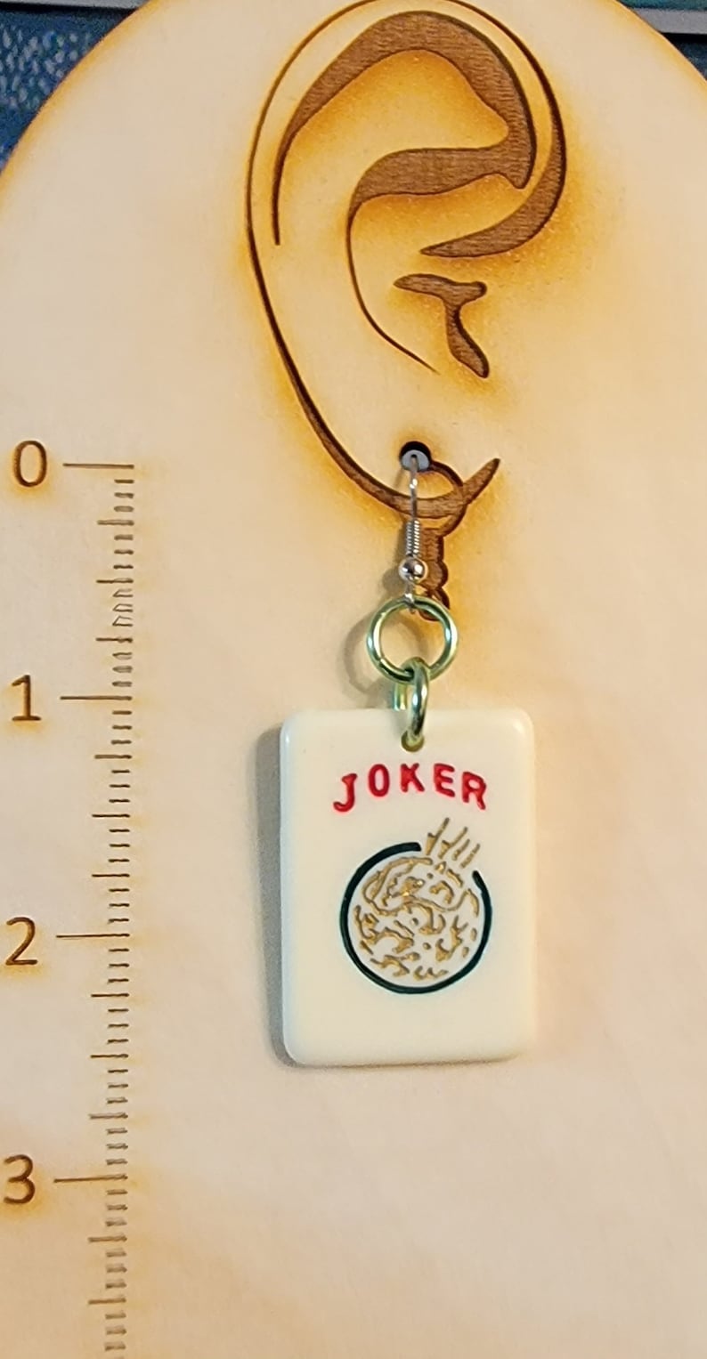 May include: A white ceramic earring with a red "JOKER" inscription and a gold-colored design. The earring is hanging from a silver hook.