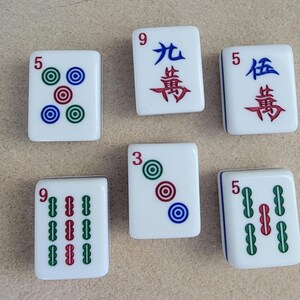May include: Six white Mahjong tiles with various markings. The tiles feature numbers, Chinese characters, and colored circles or bars. The colors include green, red, and blue. The tiles are arranged on a light brown surface.