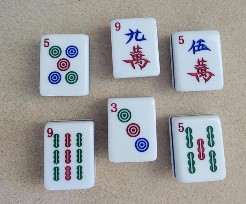 May include: Six white Mahjong tiles with various markings. The tiles feature numbers, Chinese characters, and colored circles or bars. The colors include green, red, and blue. The tiles are arranged on a light brown surface.