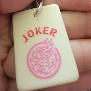 May include: A rectangular keychain with the word "JOKER" in red letters. Below the text is a pink circular design. The keychain is cream-colored and attached to a silver metal ring.