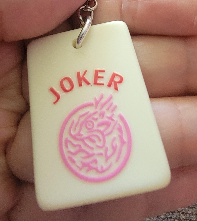 May include: A rectangular keychain with the word "JOKER" in red letters. Below the text is a pink circular design. The keychain is cream-colored and attached to a silver metal ring.