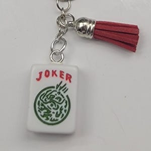 May include: A white rectangular keychain charm with the word "JOKER" in red letters. The charm has a green design and is attached to a silver chain and a red tassel.