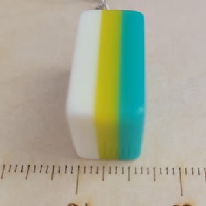 May include: A rectangular pendant with a white, yellow, and teal colour block design. The pendant is approximately 2.5 cm in length and is attached to a silver chain.