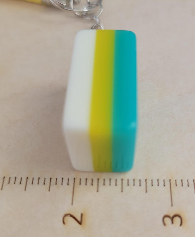May include: A rectangular pendant with a white, yellow, and teal colour block design. The pendant is approximately 2.5 cm in length and is attached to a silver chain.
