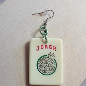 May include: A pair of white and green joker earrings with a silver hook and a green chain. The joker is a white tile with a green circle and a gold design inside. The word "JOKER" is written in red letters on the tile.