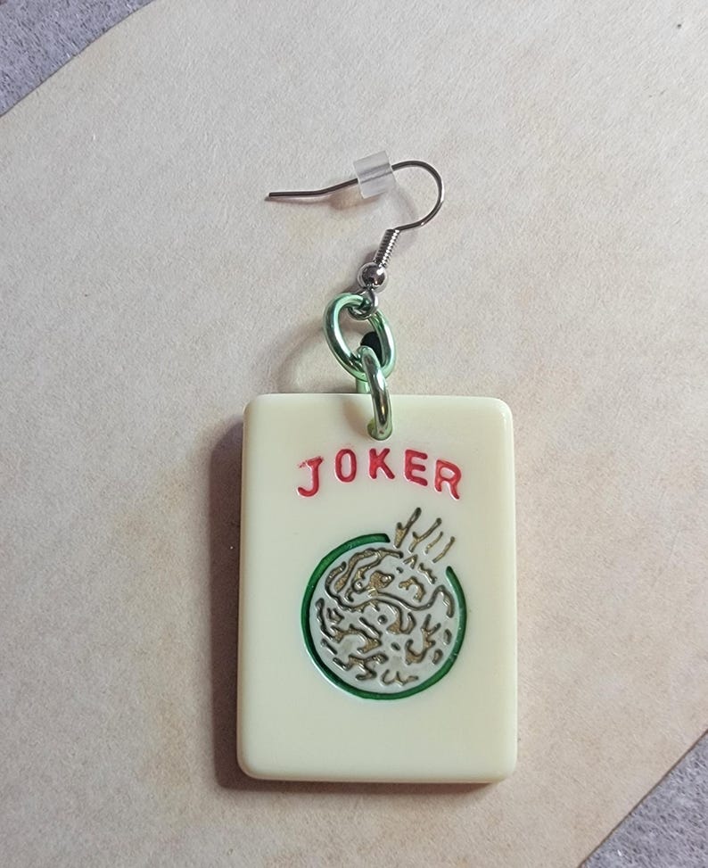 May include: A pair of white and green joker earrings with a silver hook and a green chain. The joker is a white tile with a green circle and a gold design inside. The word "JOKER" is written in red letters on the tile.