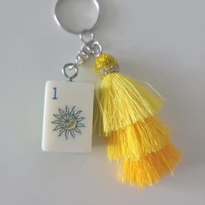 May include: A keychain featuring a white tile with the number 1 and a sun design in blue and yellow. The keychain has a silver ring and chain, and a yellow tassel with a rhinestone-studded top.