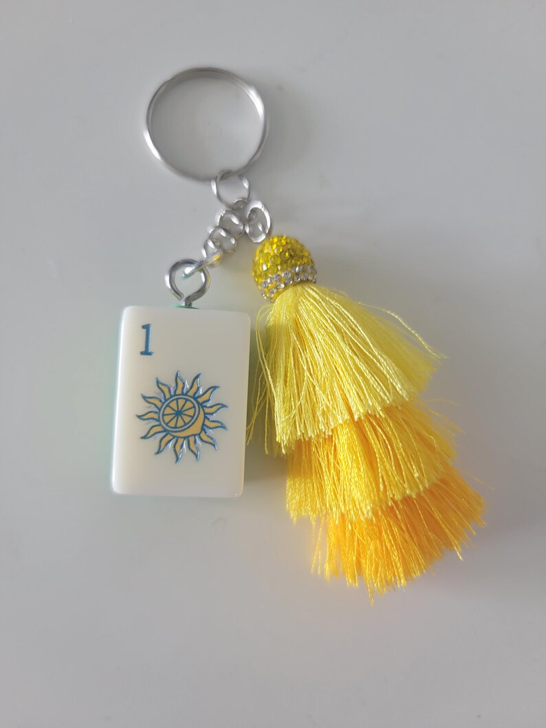 May include: A keychain featuring a white tile with the number 1 and a sun design in blue and yellow. The keychain has a silver ring and chain, and a yellow tassel with a rhinestone-studded top.