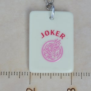 May include: A rectangular, off-white keychain charm with the word "JOKER" in red and a pink circular design. The charm is attached to a silver-colored metal keyring. The charm measures approximately 1 inch in length.