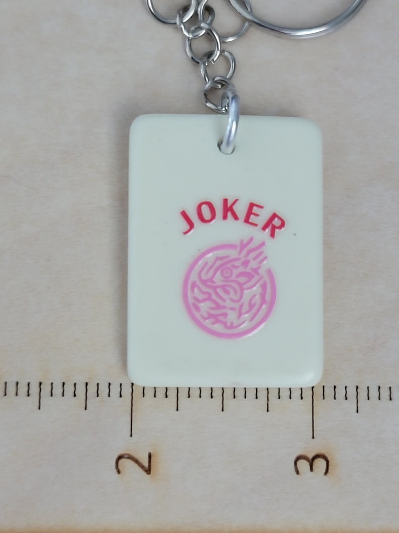 May include: A rectangular, off-white keychain charm with the word "JOKER" in red and a pink circular design. The charm is attached to a silver-colored metal keyring. The charm measures approximately 1 inch in length.