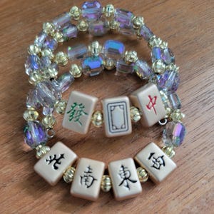 May include: Two beaded bracelets with iridescent and gold beads. The bracelets feature cream-colored Mahjong tiles with black and green Chinese characters. The bracelets are on a wooden surface.