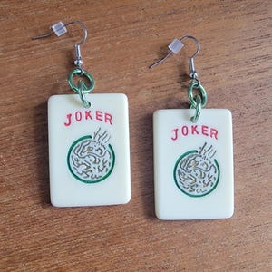 May include: A pair of earrings made from white plastic playing cards with the word "JOKER" printed in red. The cards have a green circle with a silver design in the center. The earrings are attached to silver hooks with green metal rings.