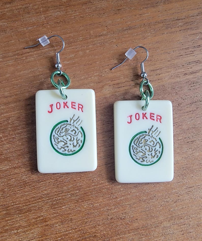 May include: A pair of earrings made from white plastic playing cards with the word "JOKER" printed in red. The cards have a green circle with a silver design in the center. The earrings are attached to silver hooks with green metal rings.