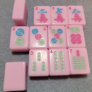 May include: A collection of pink Mahjong tiles with various symbols and numbers in blue, green, and pink. The tiles are arranged on a gray surface, showcasing the game's components.